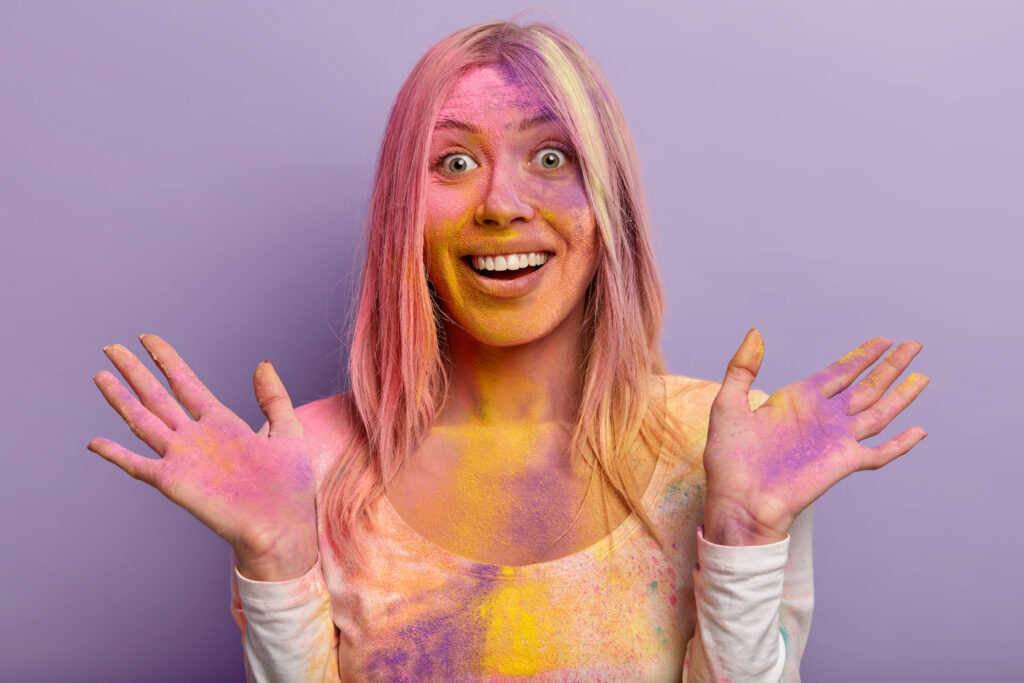 Festivals of Facial: Get Your Skin Holi-Ready with the Perfect Pre-Glow Ritual 2 headshot glad female with toothy smile happy reaction spreads hands dirty with multicolored dry powder has fun traditional holi festival isolated against purple wall vibrant colors